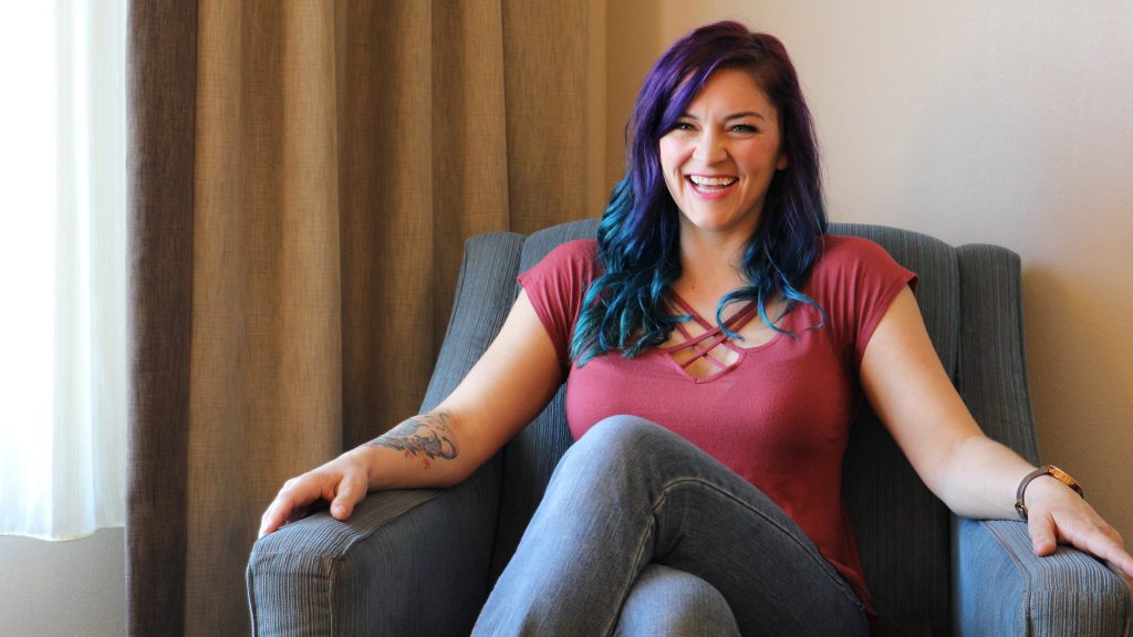 A young woman with purple and blue hair sits in a blue armchair as she laughs while telling a story.